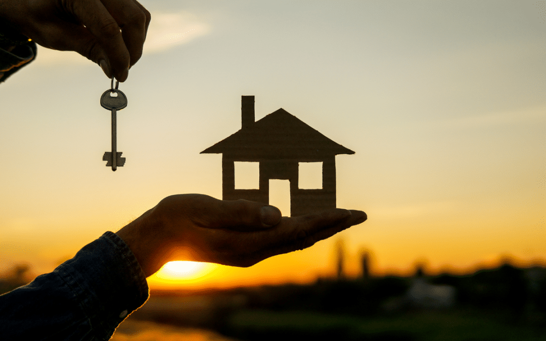 Hand holding a house model and a key against a sunset background