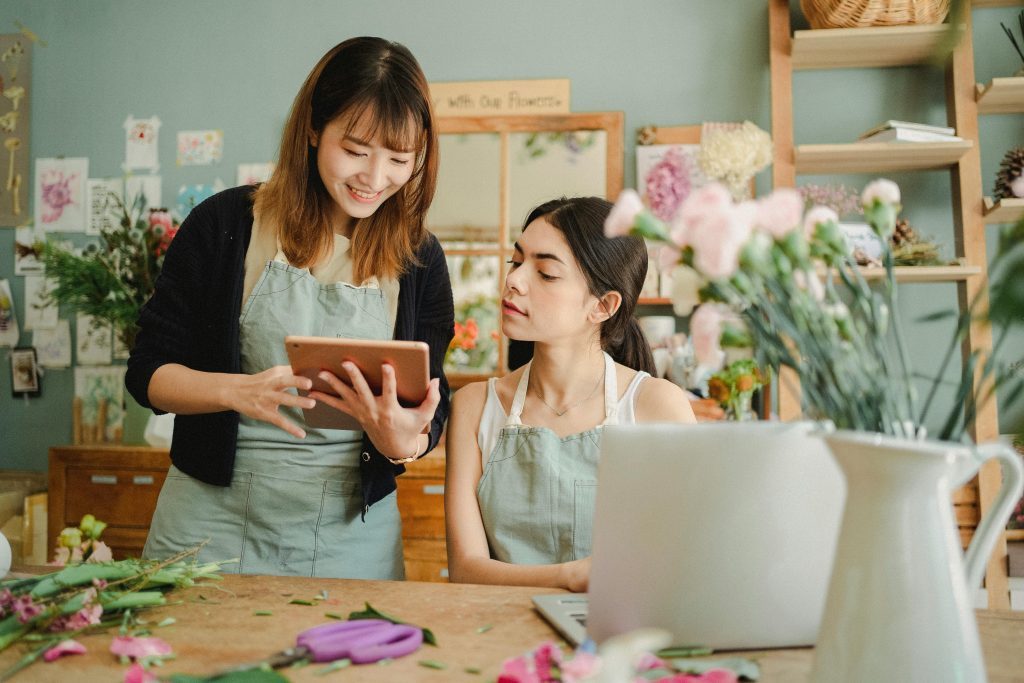 Two women in aprons working in a cozy flower shop with a tablet and laptop surrounded by fresh flowers and plants