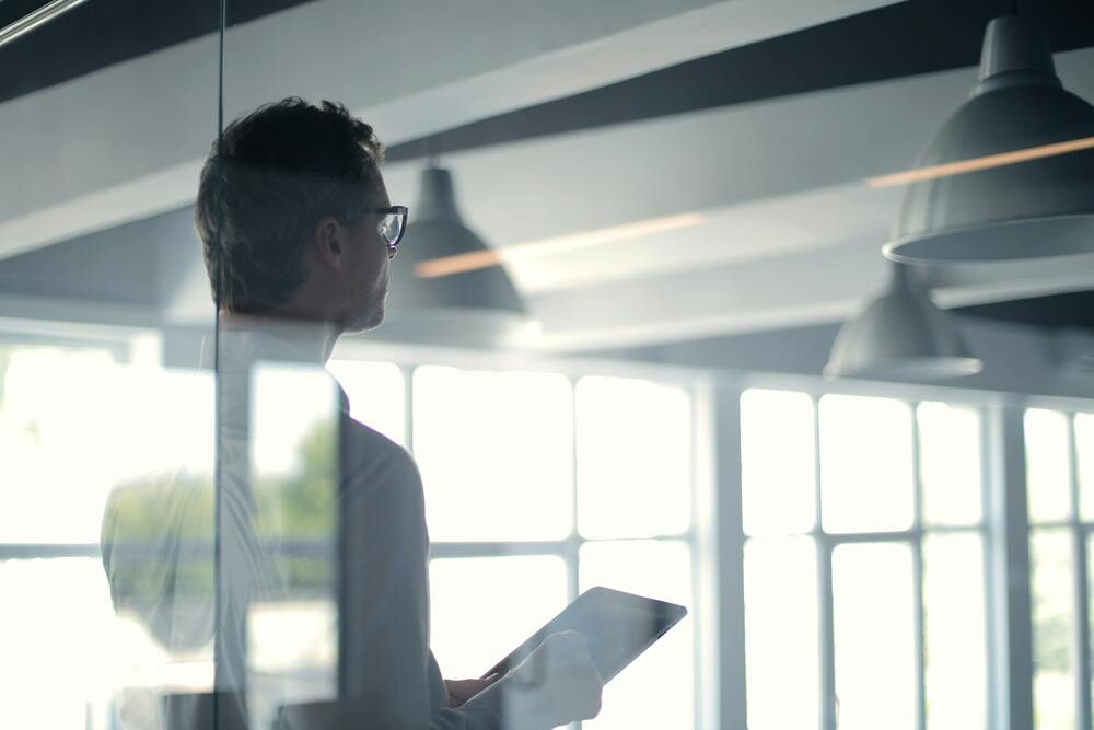 Businessman with tablet in modern office, looking through large windows.