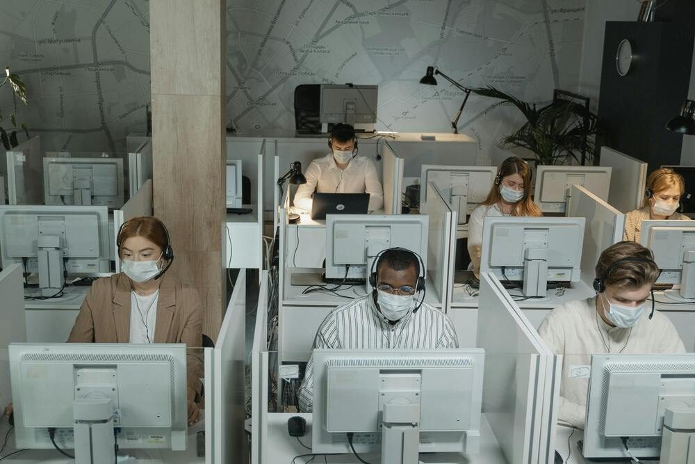 Diverse call center employees wearing masks and working on computers in cubicles.