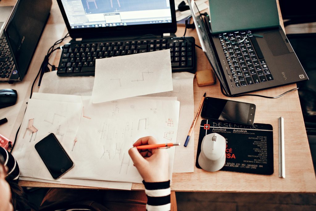 Designer working on architectural sketches with laptops, smartphones, and drawing tools on desk.