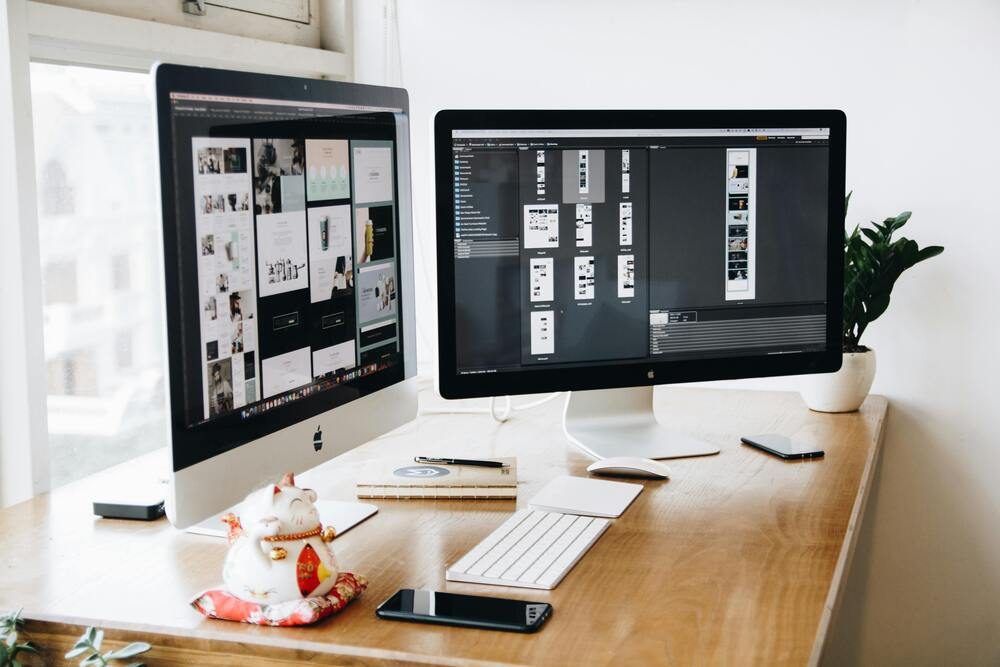 Dual screen computer setup for graphic design work on a wooden desk.