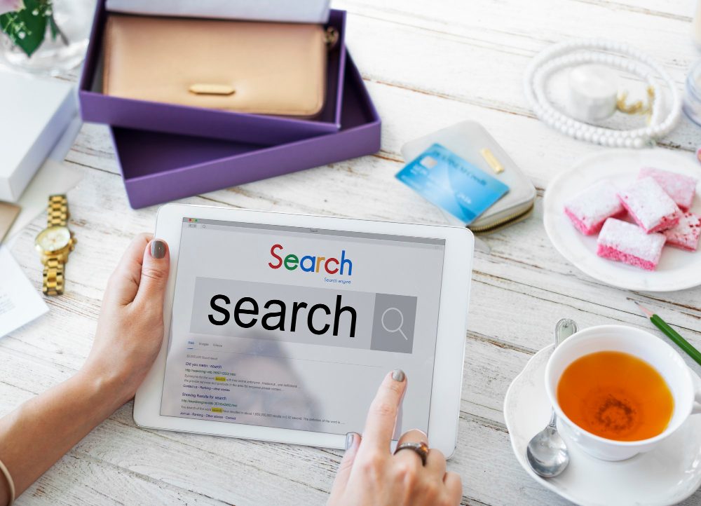 Person using a tablet with the word search displayed on the screen, surrounded by a wallet, credit card, tea, and pastries.