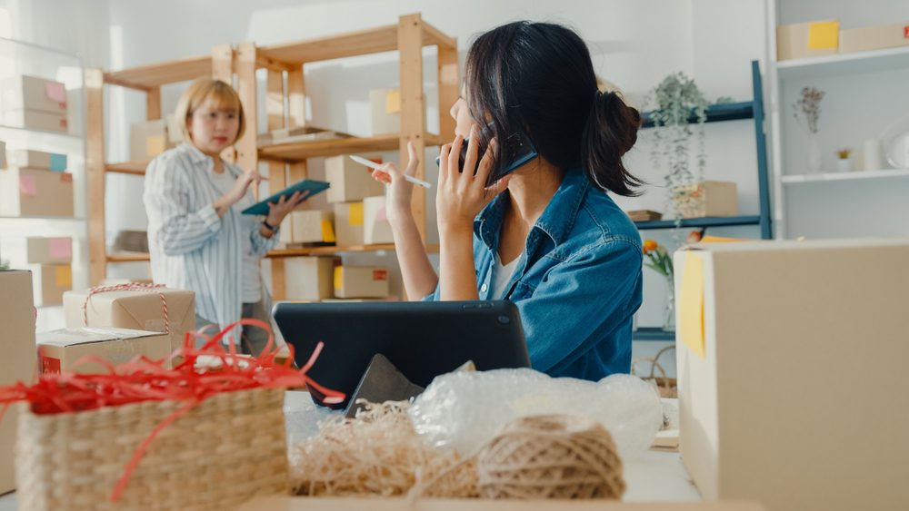 Two women organizing a small business workspace surrounded by boxes and packaging materials.