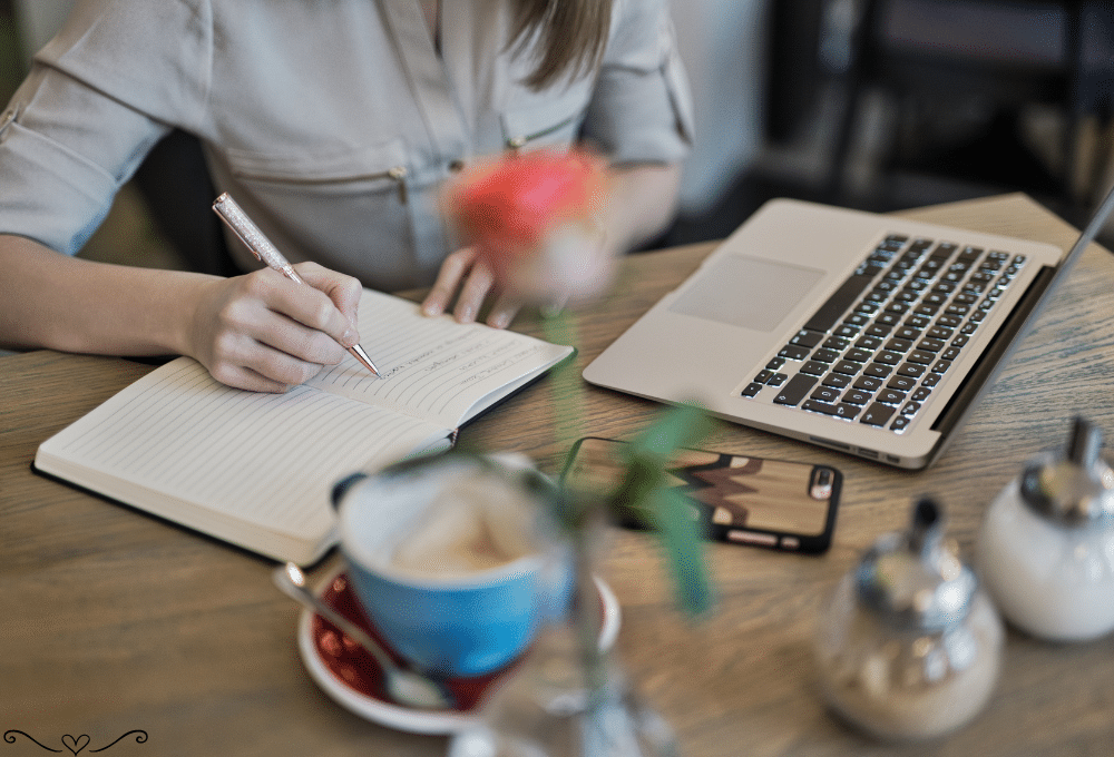 Person writing in a notebook at a café table with a laptop, phone, and coffee.