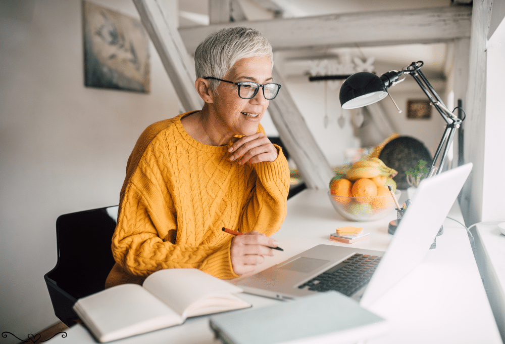 Smiling senior woman in a yellow sweater working on a laptop at a bright home office desk.