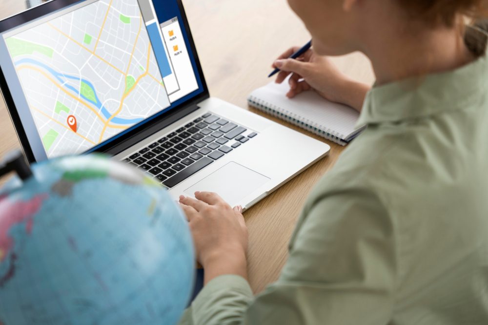 Person using a laptop with a map on the screen and a globe nearby, taking notes at a desk.