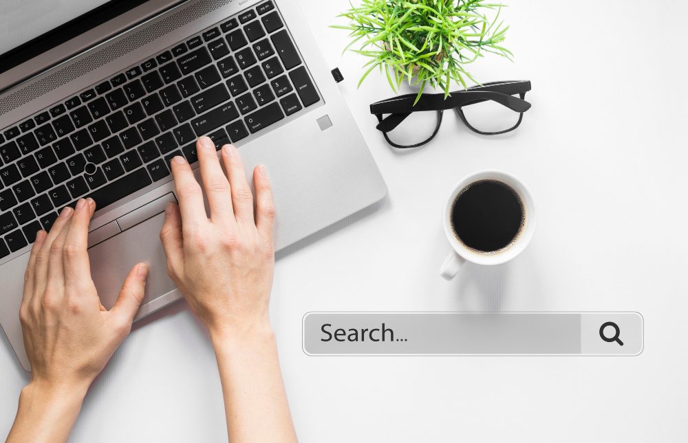 Hands typing on a laptop keyboard beside a coffee cup, glasses, and plant.