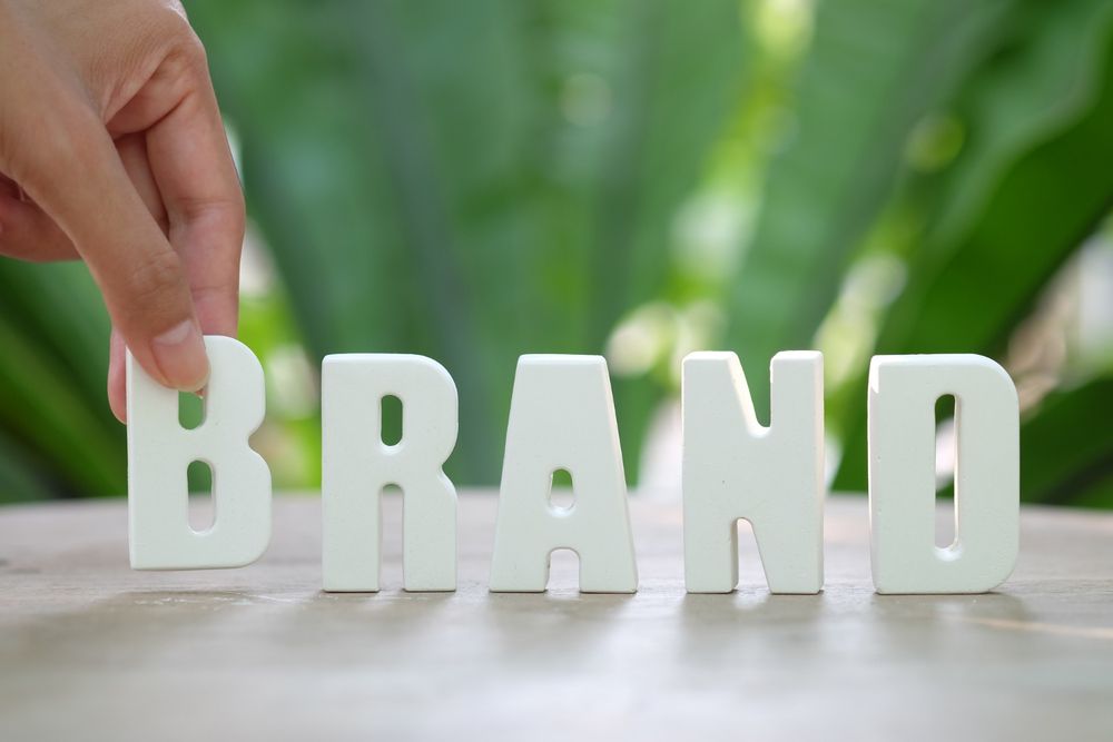 Hand arranging white letters spelling "BRAND" on a table with green foliage background